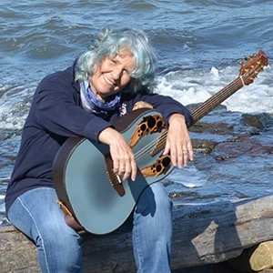 Lucie Blue Tremblay by the rocky seashore holding her guitar